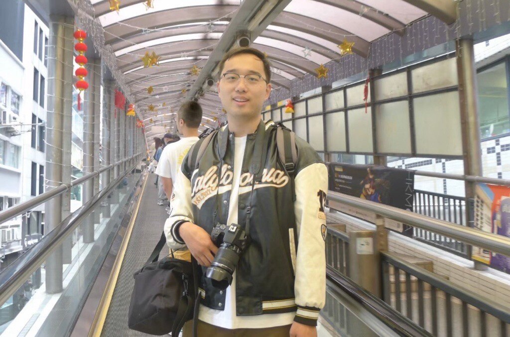 Pipi Tang smiles as he holds a small bag and a camera while posing for a photo in a covered walkway.