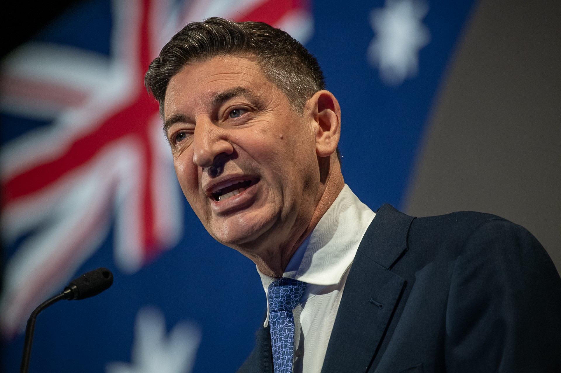 A man with dark hair and a dark suit speaks with the Australian flag behind him