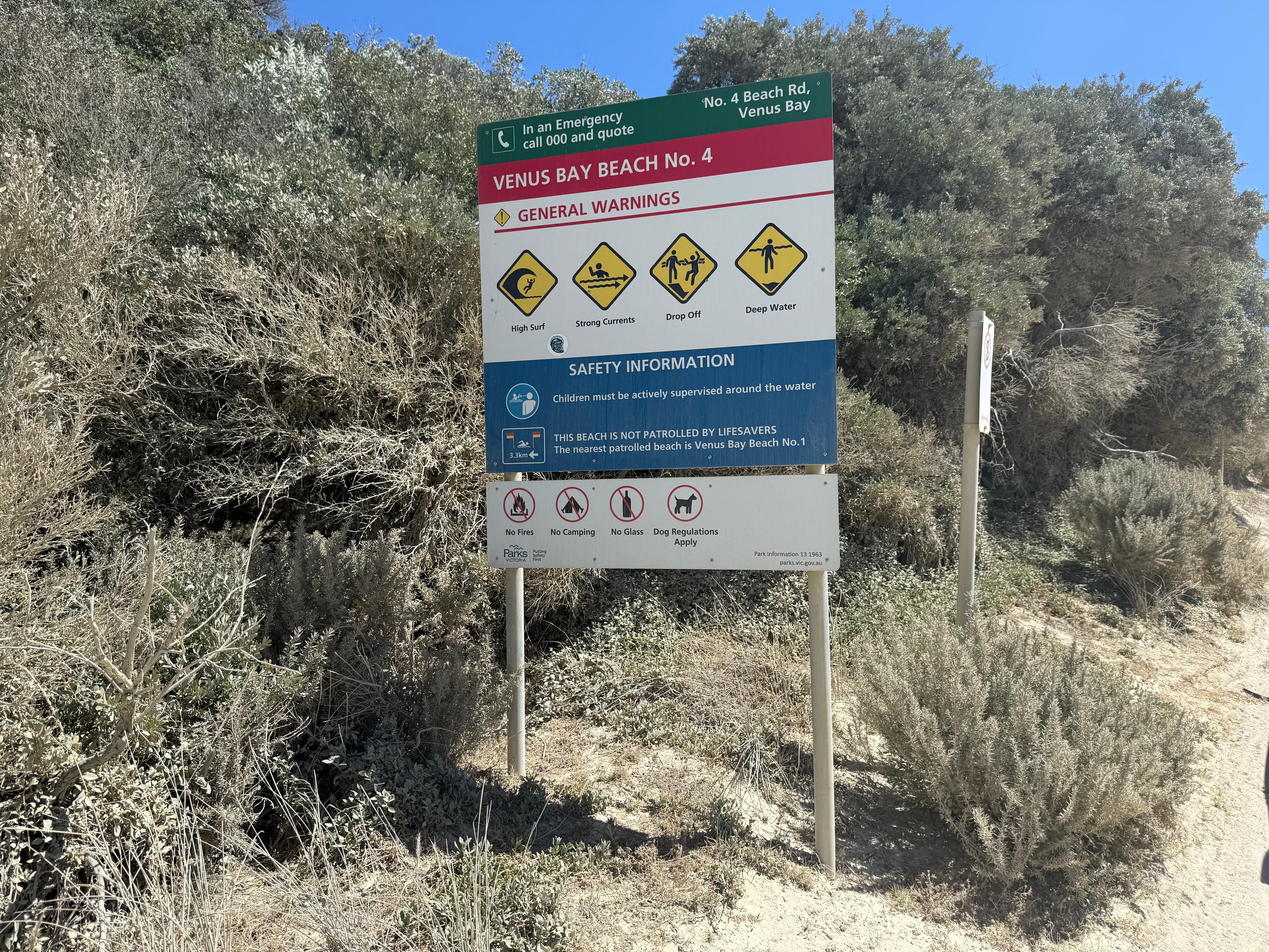 Uma foto de um sinal de alerta de praia preto, verde, vermelho, branco, amarelo e azul, situado em frente a arbustos verdes na praia.