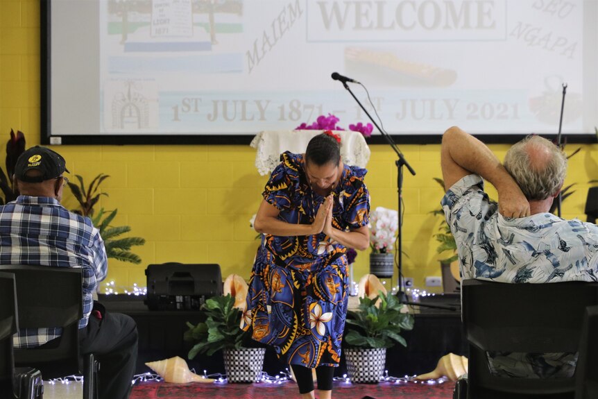 An Islander woman wearing a dark blue patterned frock bows with her hands together, as if in prayer 