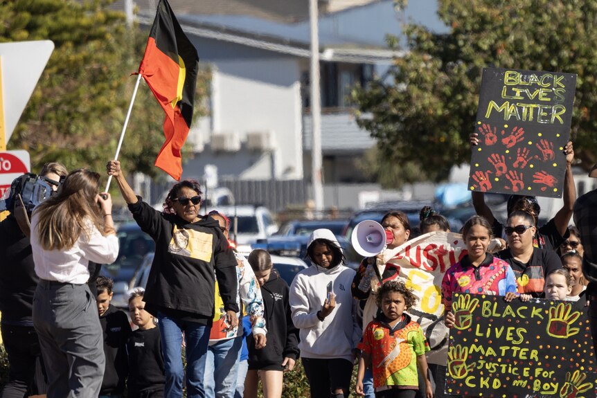 Dozens of people walk through the street in Geraldton holding placards and Aboriginal flags.