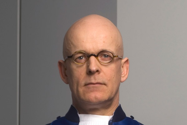A bald man wearing glasses and a Judge's robes looks at the camera with a serious expression