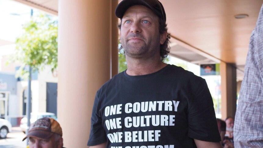 A man stands in a black shirt with the words 'One country one culture one belief one custom'.