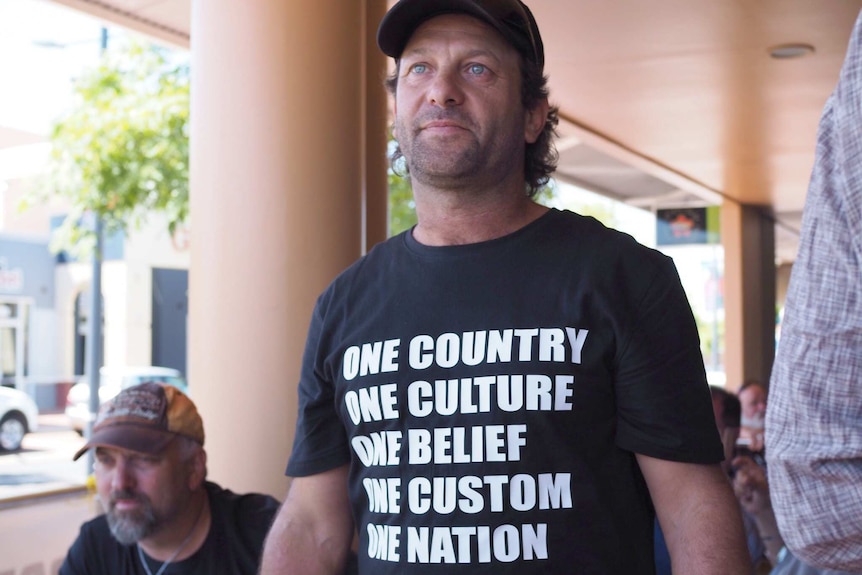 A man stands in a black shirt wih the words 'One country one culture one belief one custom'.