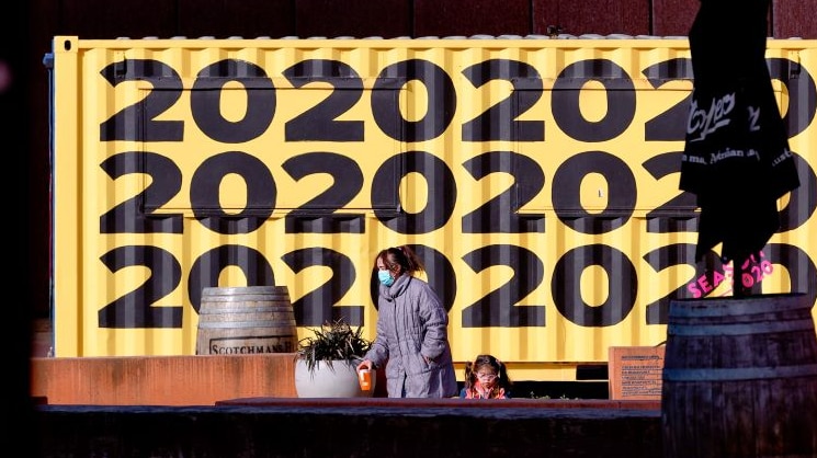 A woman wearing a mask and a grey winter coat stands beside a young girl in front of a yellow mural with '2020' written on it