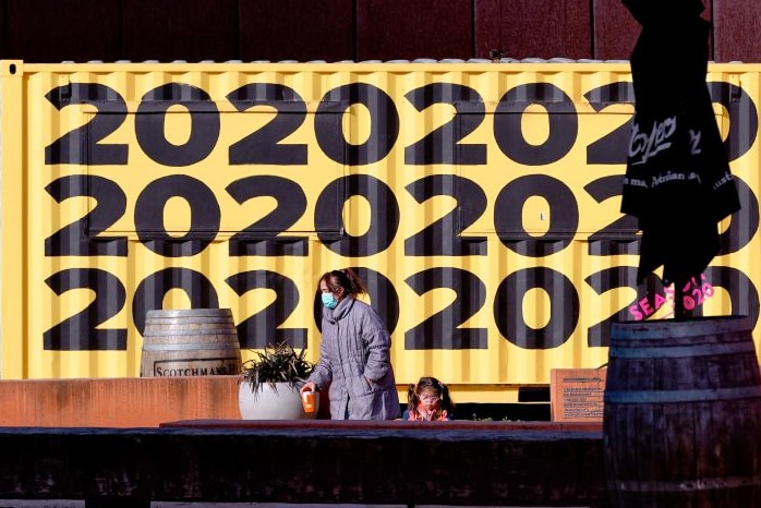 A woman wearing a mask and a grey winter coat stands beside a young girl in front of a yellow mural with '2020' written on it