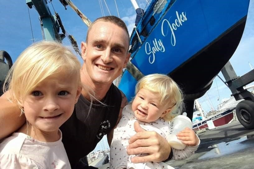 Man hold two young girls in front of a boat named Salty Jocks.