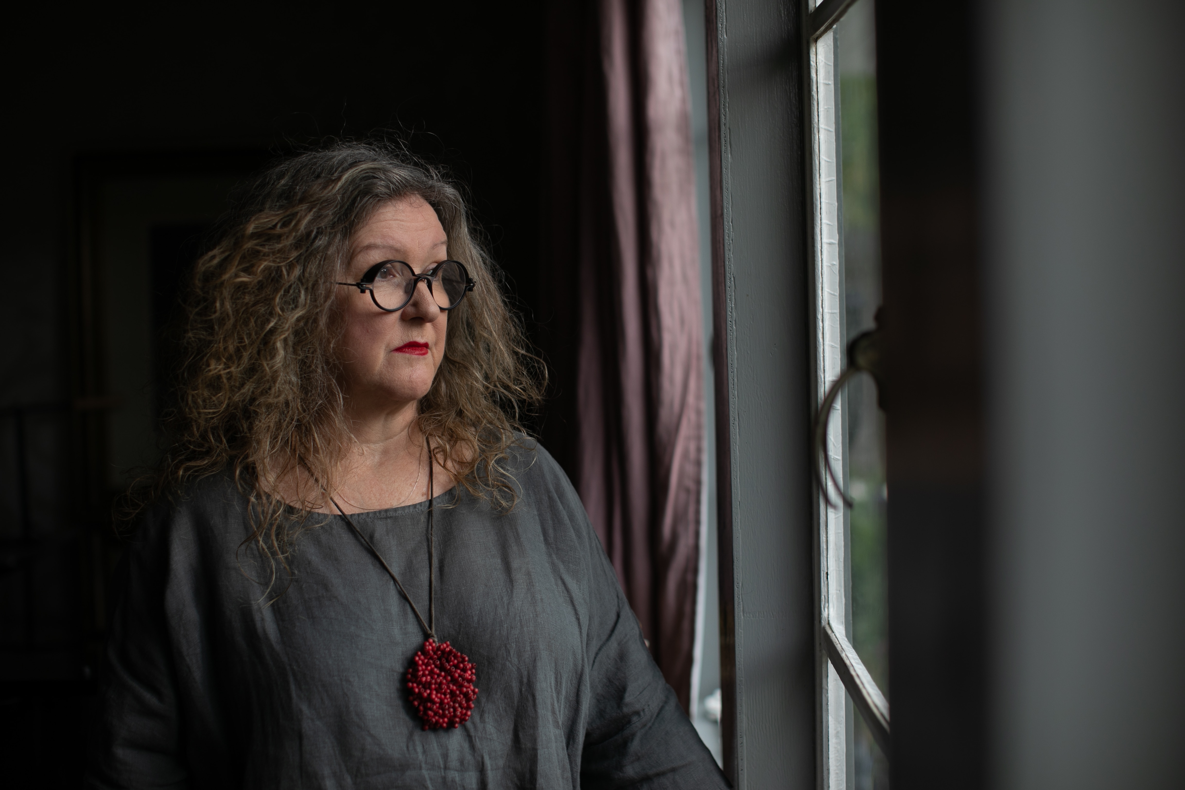 A woman wearing loose grey linen top with red necklace gazes out of a window