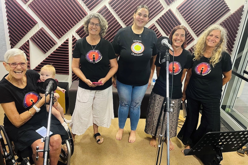 Five smiling women wearing tee-shirts with Indigenous logo stand in front of mics, elderly woman holds baby.