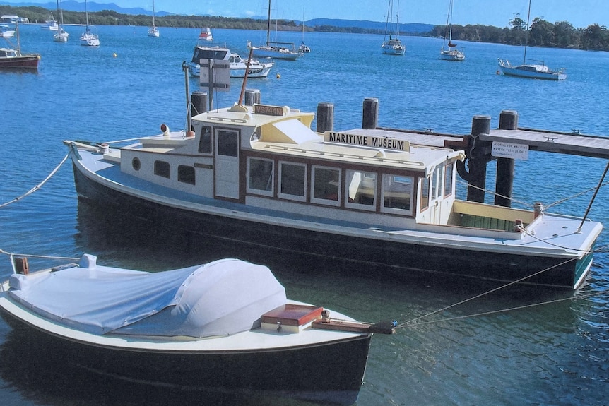 A restored wooden vessel, on a mooring on a river, with other boats in the background.