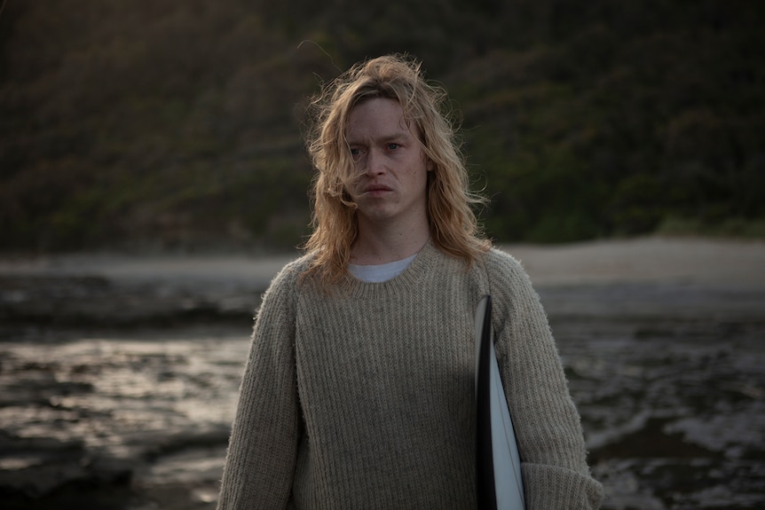 Young man with wavy blonde hair stands on a beach wearing a taupe knitted jumper, holding a surfboard and looking disgruntled.