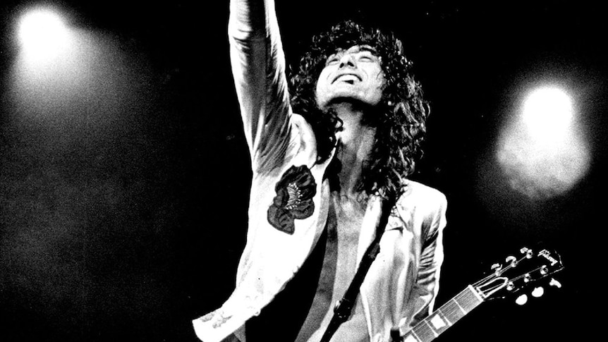 Jimmy Page with electric guitar kneeling upright on stage with arm pointed to sky
