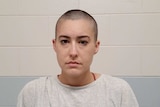 A woman with a shaved head looking at the camera, with a brick wall behind her.
