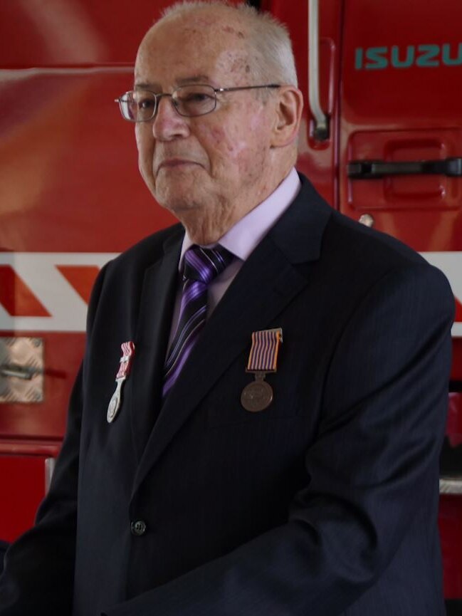 An elderly firefighter wearing a suit stands at a lectern in front of a red fire truck