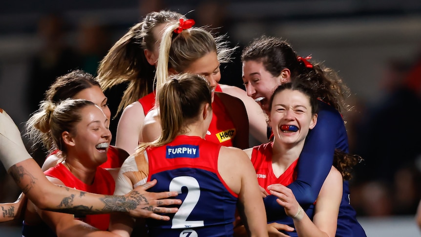 A grinning Melbourne AFLW player is surrounded and hugged by her teammates after kicking a goal.