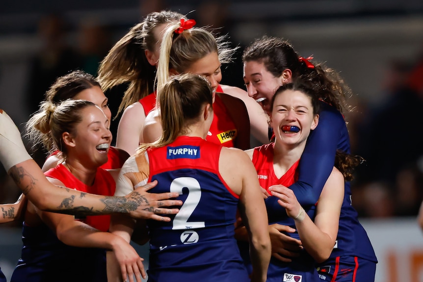 A grinning Melbourne AFLW player is surrounded and hugged by her teammates after kicking a goal.