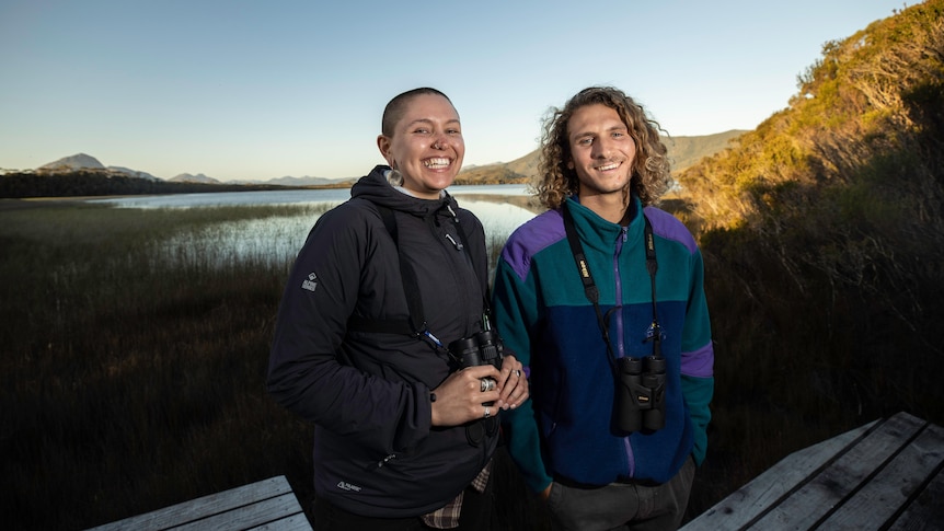 A woman and a man each with a pair of binoculars around their neck stand in front of a lagoon at dusk and smile at the camera
