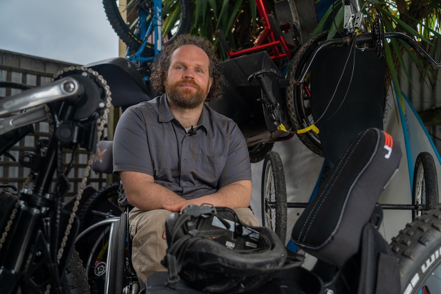 a man sits in a wheel chair in a shed with many adaptive bikes hanging behind him.