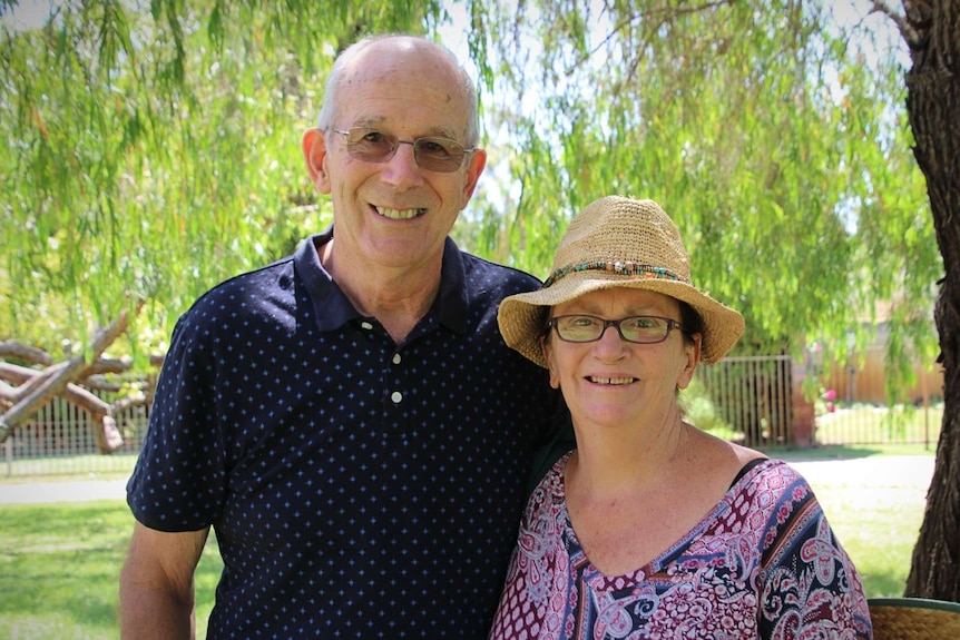An older couple stand amongst the shade of a tree in a park