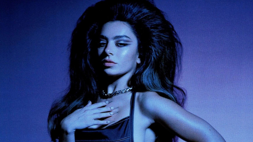 A blue toned image of Charli XCX who wears a black mini dress and big teased hair. She smizes and has her hand on her chest.