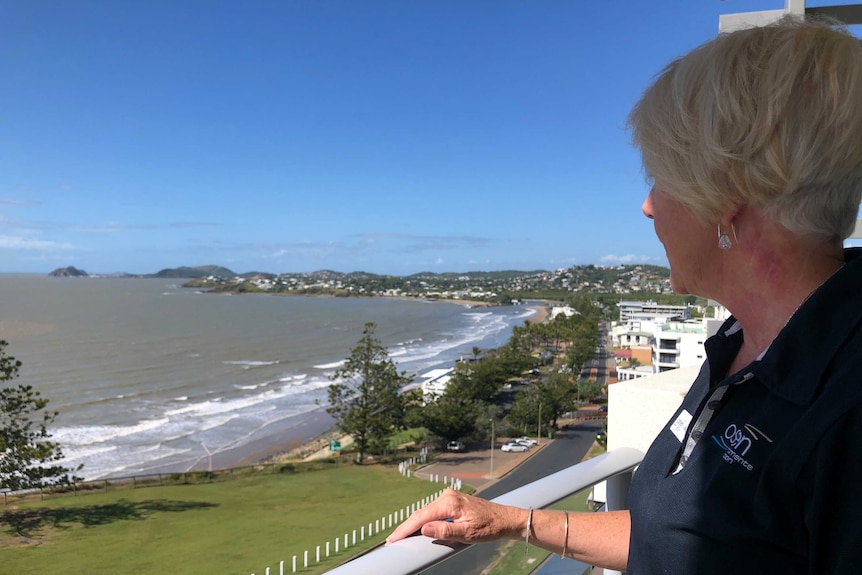 A woman looks out over a balcony at the ocean and small beach town.