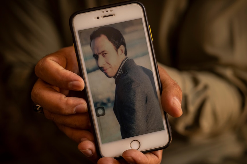 A man holds out his phone screen to display an image of a man with short dark hair 