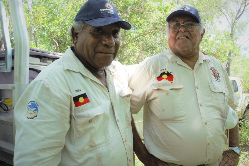 Two Indigenous men in ranger uniforms stand together in the bush.