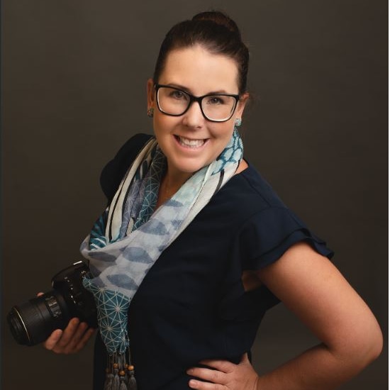 Amy Quist, a woman wearing spectacles and a scarf, stands with one hand on hip, holding a camera.