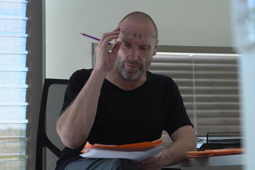 A man sitting at a desk, reading papers, with one hand lifting his glasses up so they are off his eyes