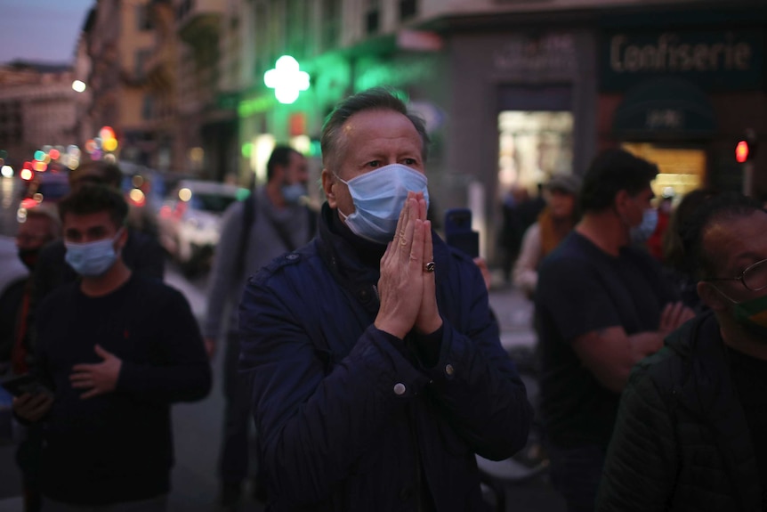 A man wearing a facemask standing in the street with his hands up in a prayer sign.