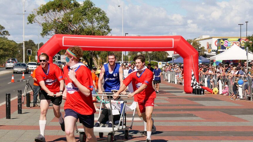 four people running and pushing a purpose built 'hospital bed' one person sits down holding the metal bar while spectators watch