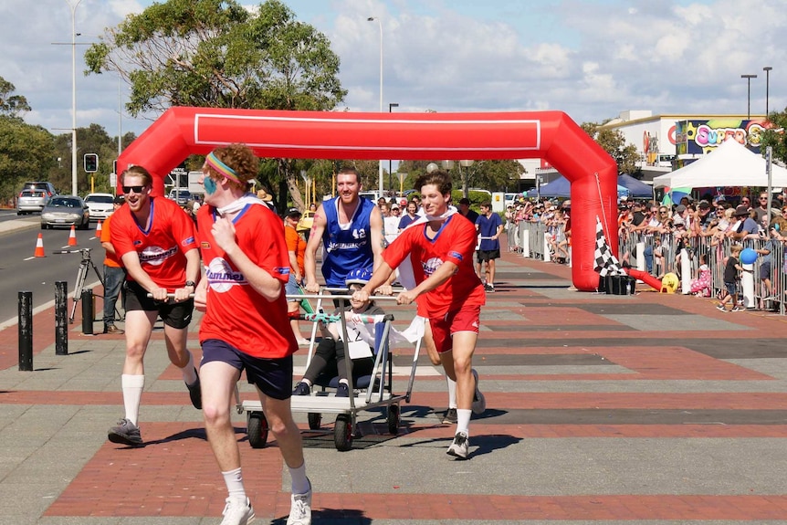 four people running and pushing a purpose built 'hospital bed' one person sits down holding the metal bar while spectators watch