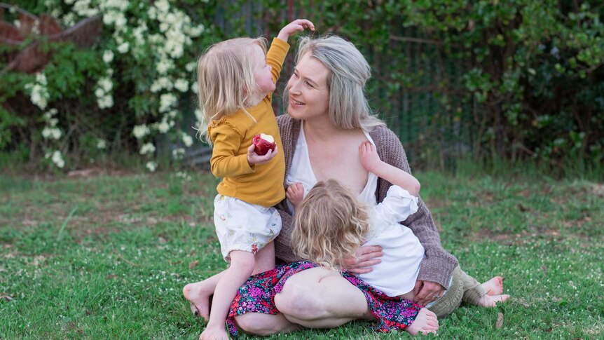A woman with blonde hair and two toddlers sitting on the grass with one of the toddlers breastfeeding.