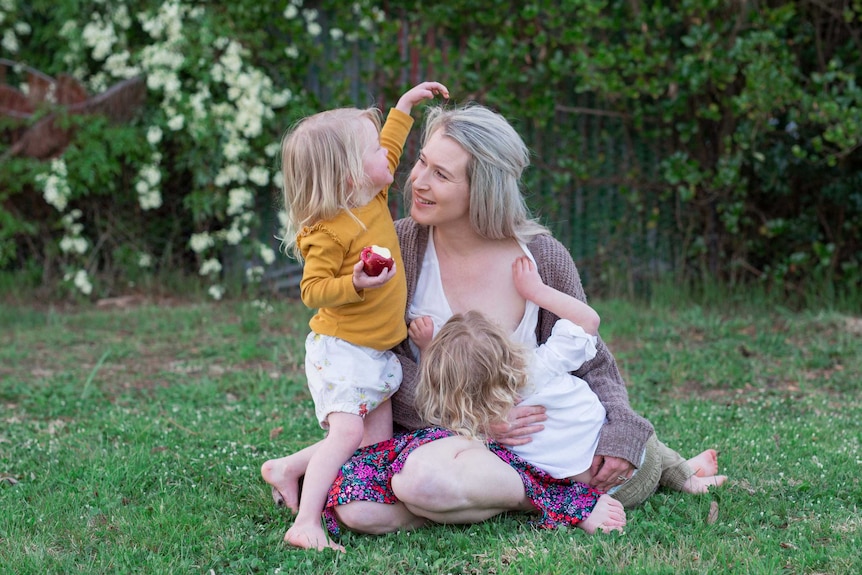 A woman with blonde hair and two toddlers sitting on the grass with one of the toddlers breastfeeding.