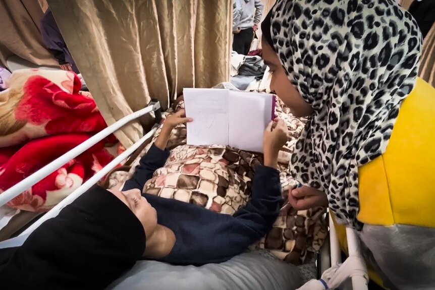 A girl lying on a hospital bed flips through a book while someone looks over her shoulder.