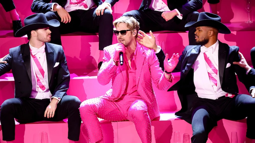 Ryan Gosling wears a bright pink suit and is seated and holding a microphone
