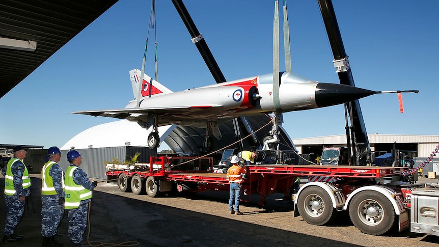 A crane lifts up a large aircraft, three men with hard hats look on