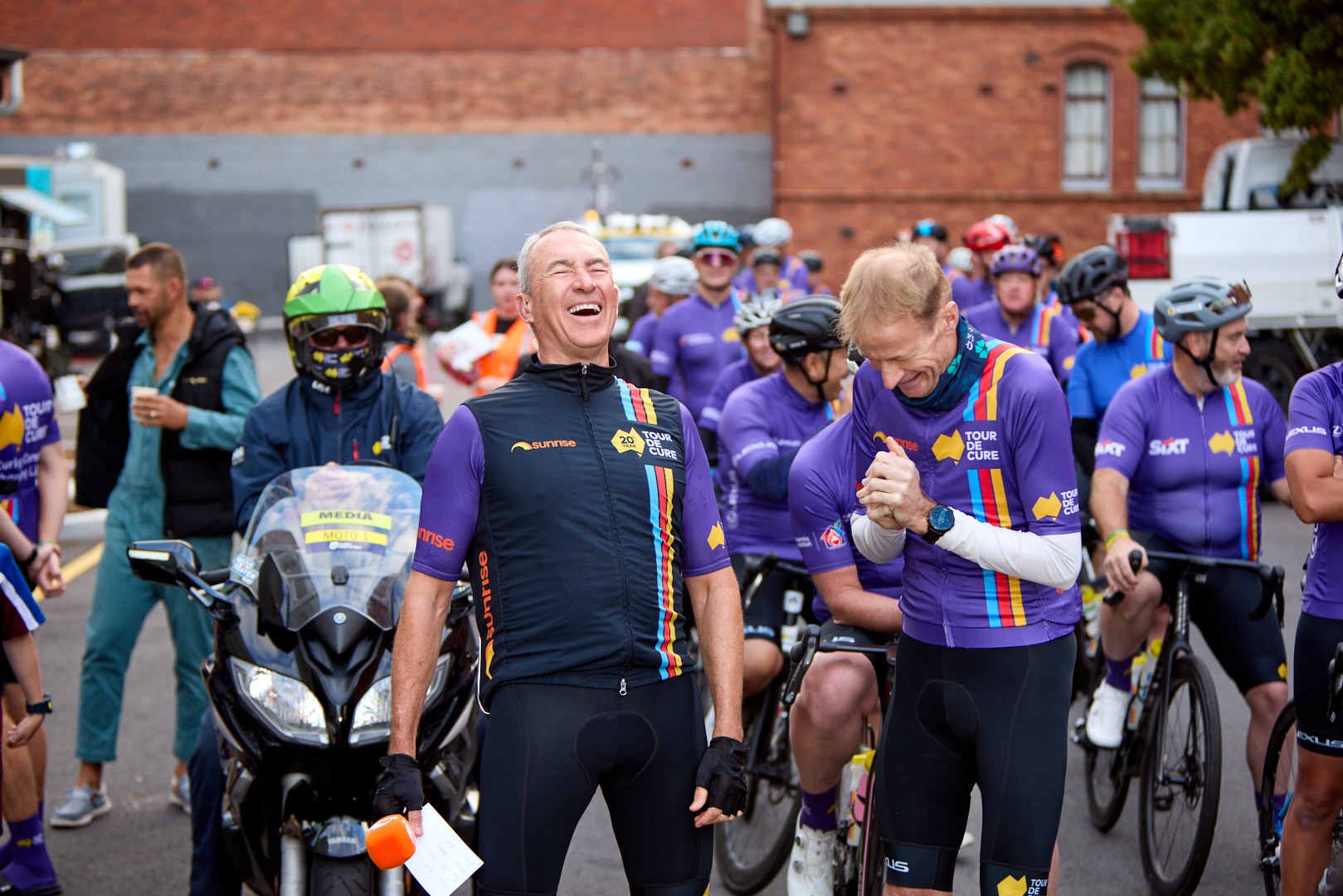 Mark Beretta and Richard Scolyer smiling and laughing, with fellow bike riders in lyra behind them.