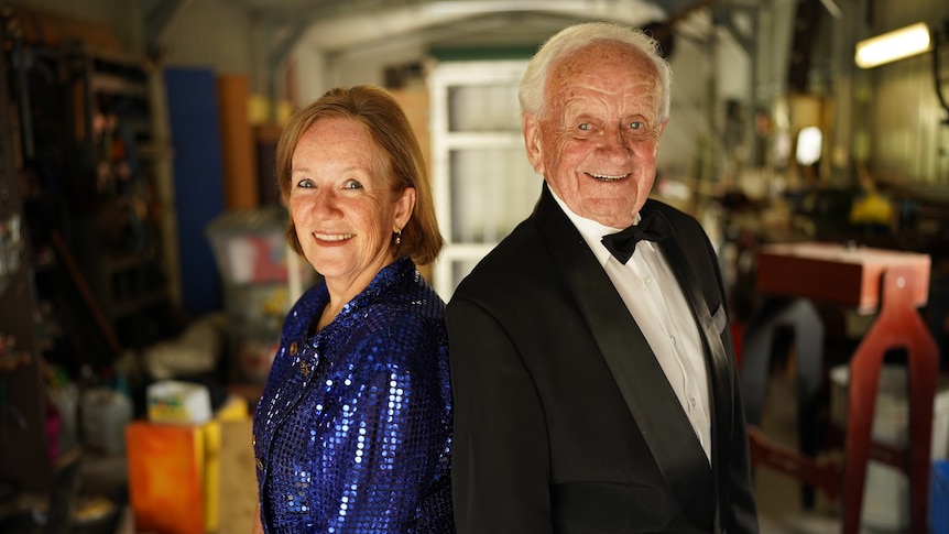 A woman in a sequin dress and an elderly man in black suit and bowtie stand back-to-back