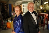 A woman in a sequin dress and an elderly man in black suit and bowtie stand back-to-back