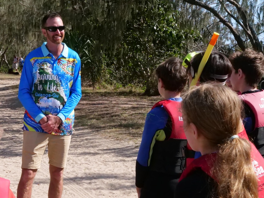 A man stands on a sand path and speaks to a group of children wearing wetsuits and snorkels