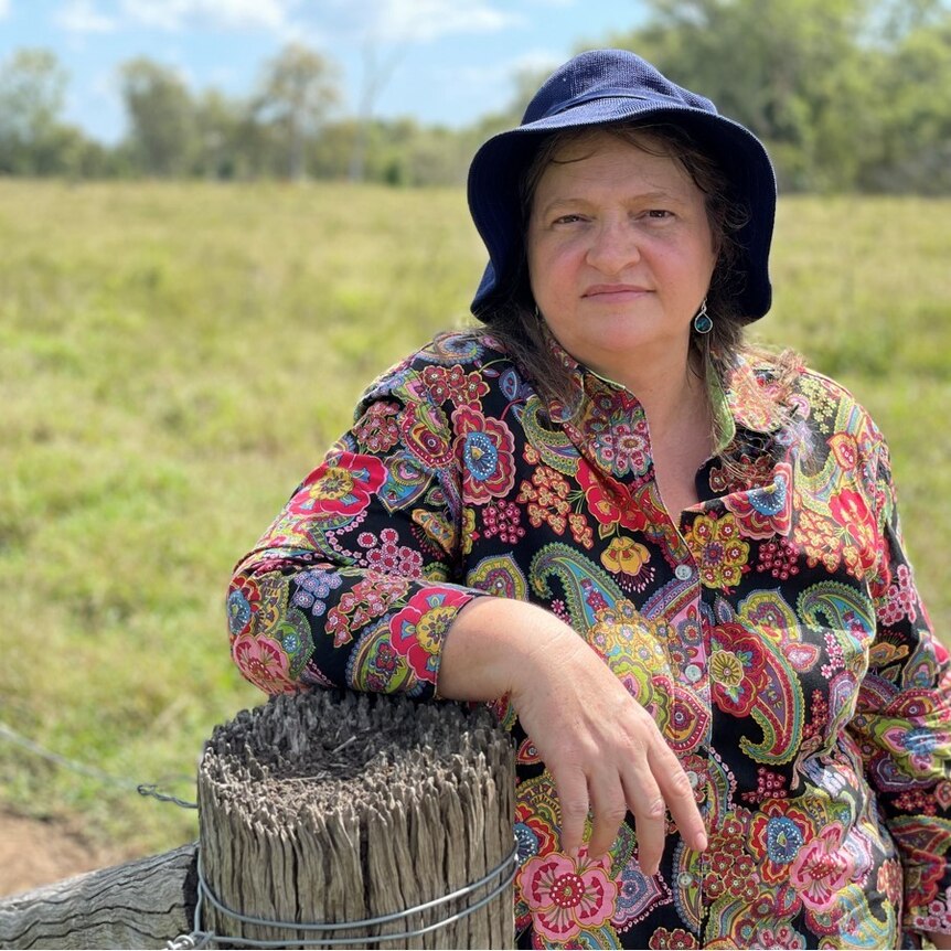 A woman stands in a paddock, leaning on a fence post wearing a colourful shirt and a blue hat
