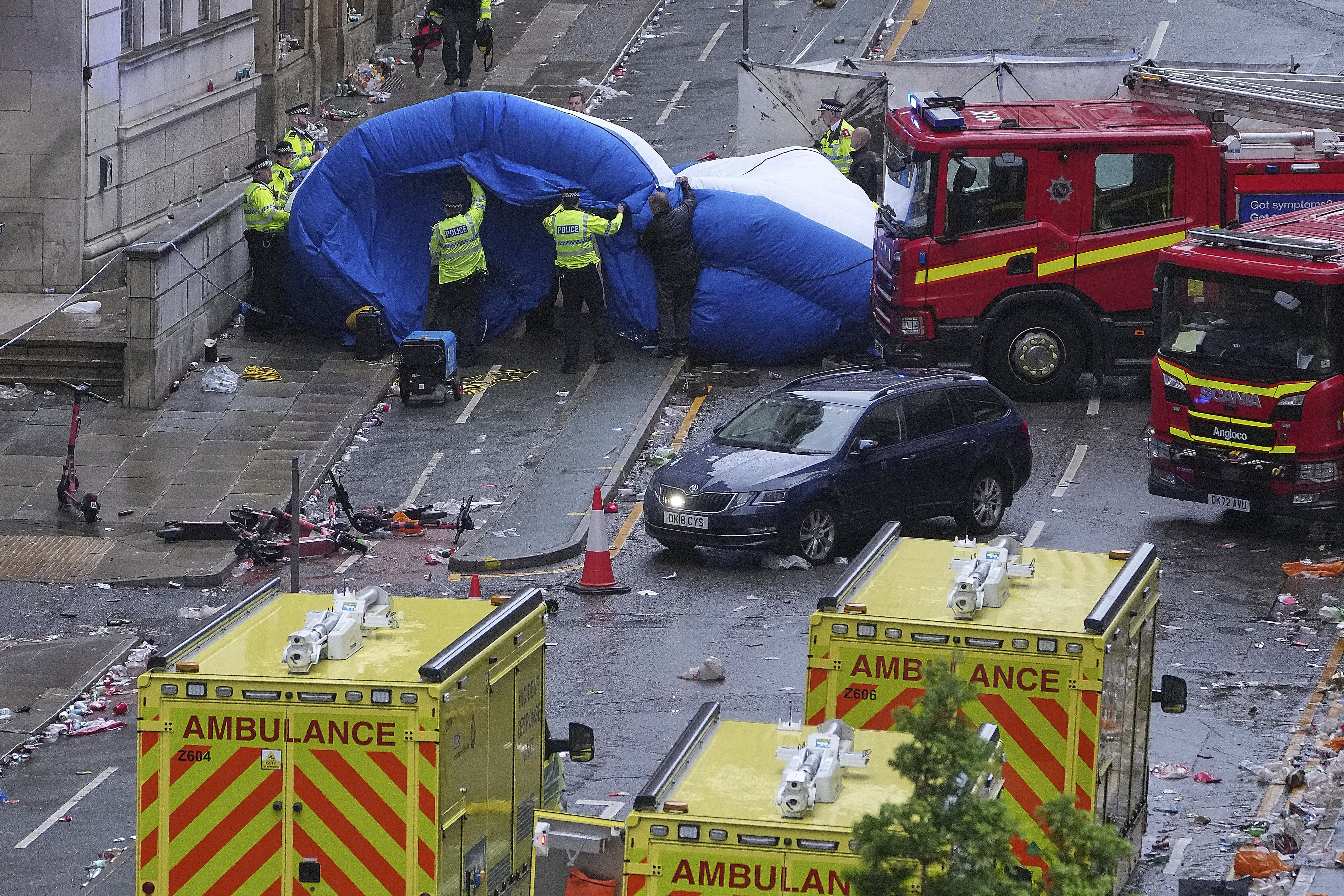 Police erect a screen as fire trucks and ambulances line a street amid debris