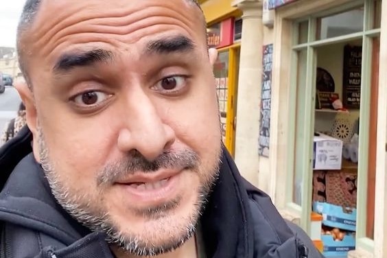 An Indian man in a black puffer jacket talks to the camera on a London street