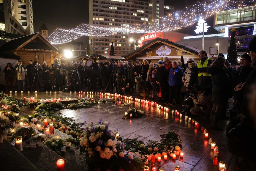 A crowd of people gather at a memorial site in Berlin, standing behind an arch lined with candles.