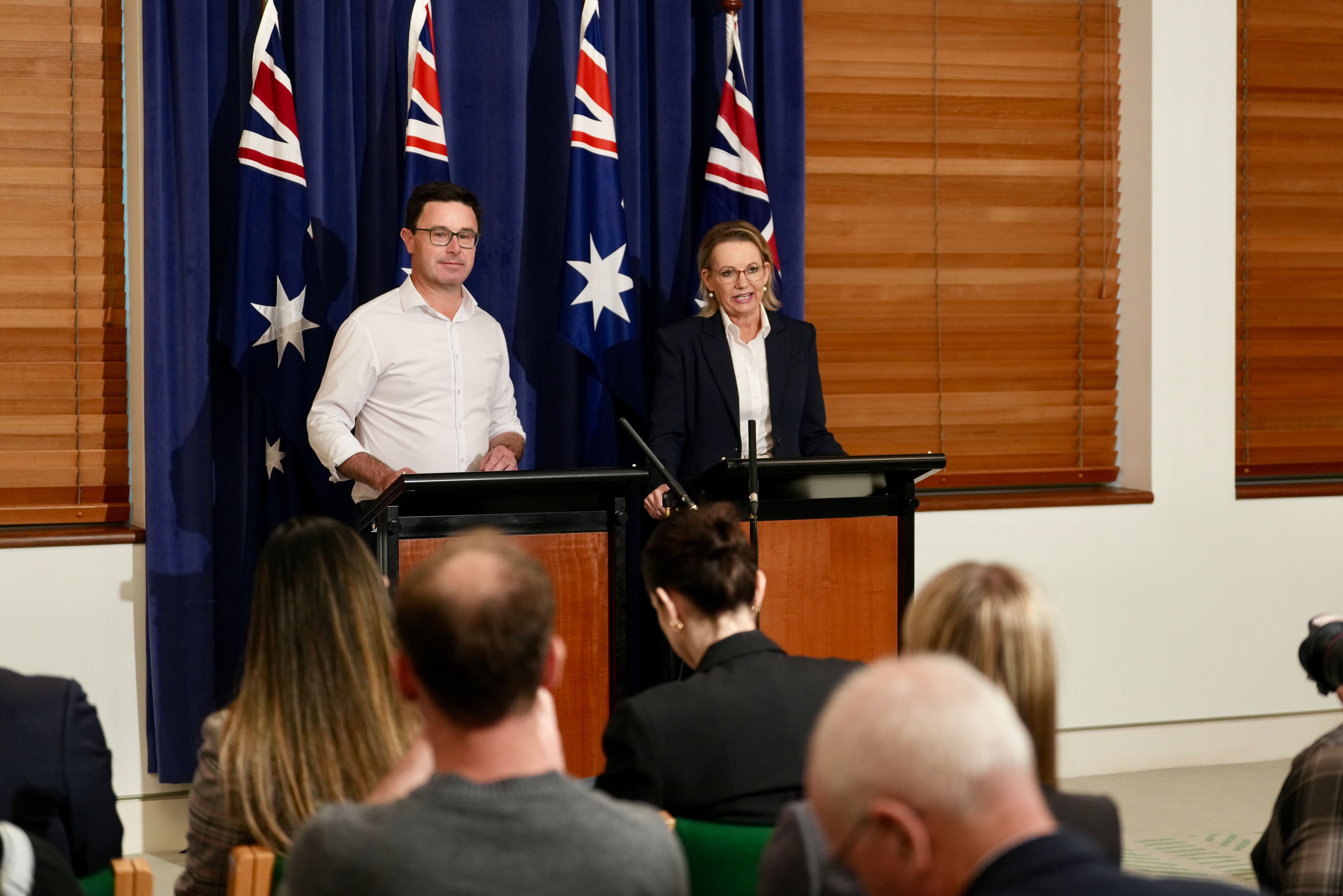 একজন পুরুষ এবং একজন মহিলা অস্ট্রেলিয়ান পতাকার পটভূমিতে একটি মঞ্চে দাঁড়িয়ে সাংবাদিকদের একটি দলের সাথে কথা বলছেন।