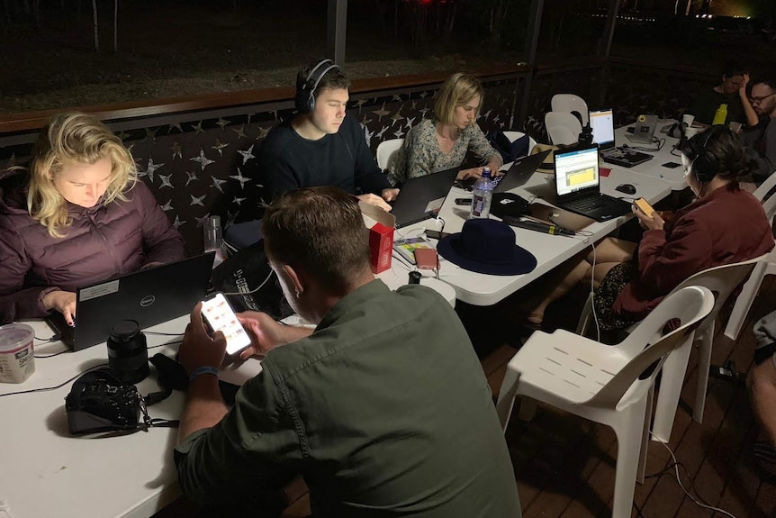 Six journalists sitting at tables working on laptops in dark.