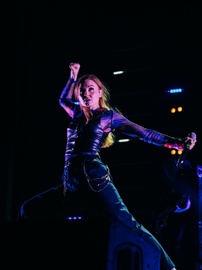 A woman strikes a pose on stage, one fist int he air and the other holding a microphone. She wears a leather vest and blue pants
