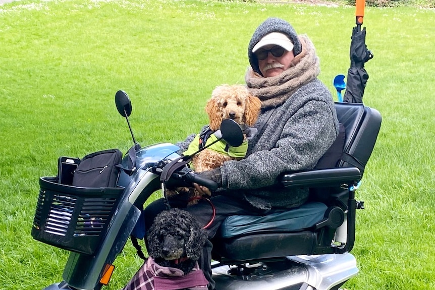  A man in a hoodie and scarf sits on a mobility scooter with a dog on his lap