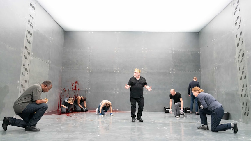 In a grey-walled room, seven people surround and kneel towards Simon Russell Beale wearing a gold crown.